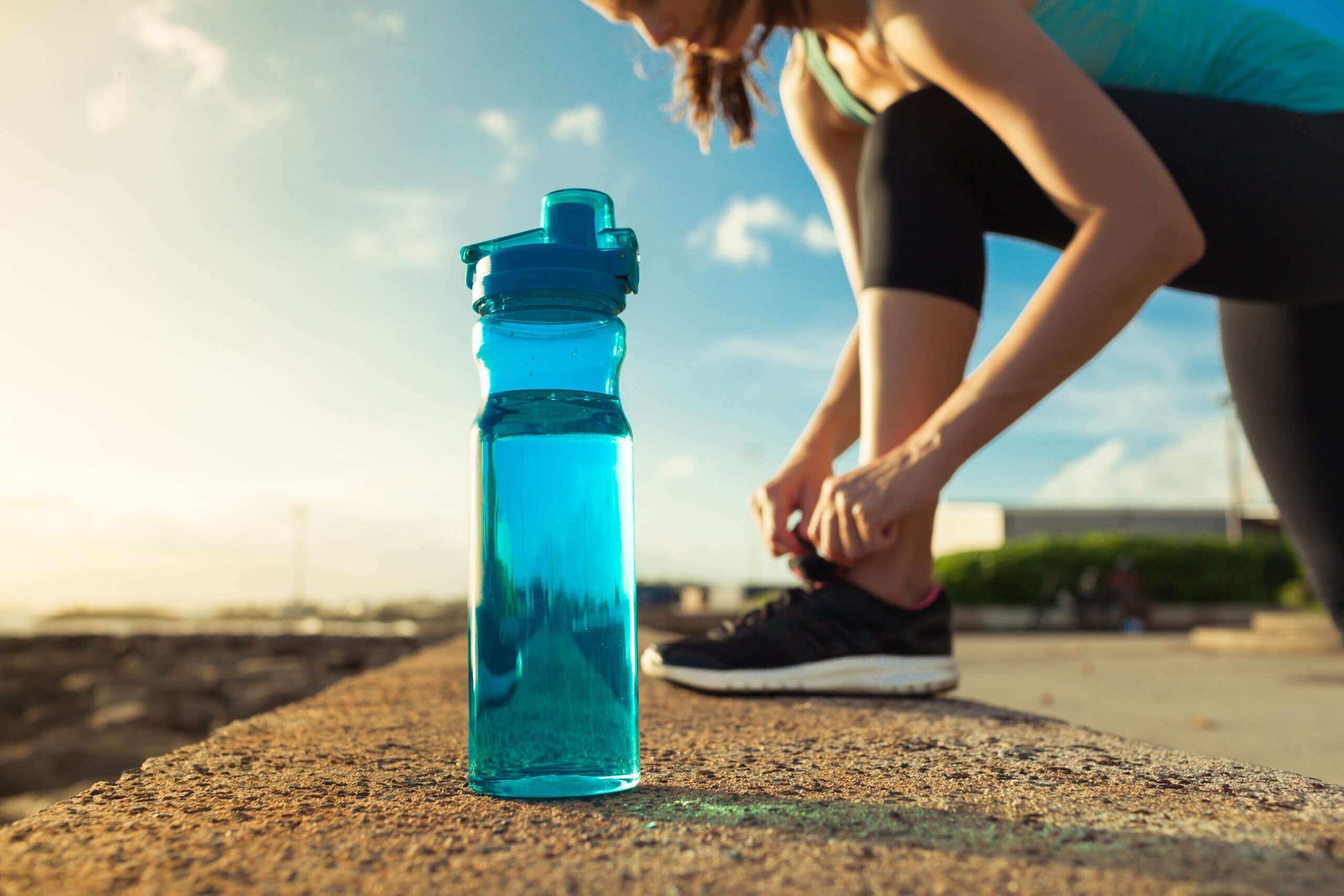A close-up of a blue water bottle on a ledge with a person in athletic wear tying their shoe in the background, outdoors in the sunlight.