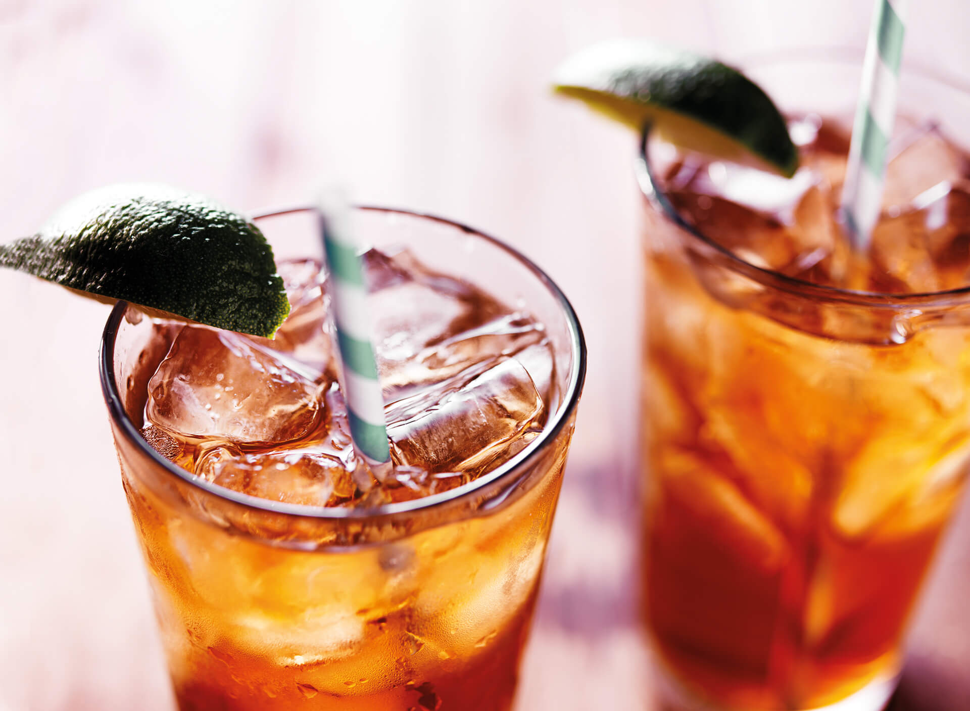 Two glasses of iced tea with striped straws, filled with ice cubes and garnished with lime wedges, invite you into the world of taste as they sit on a wooden surface bathed in warm light.