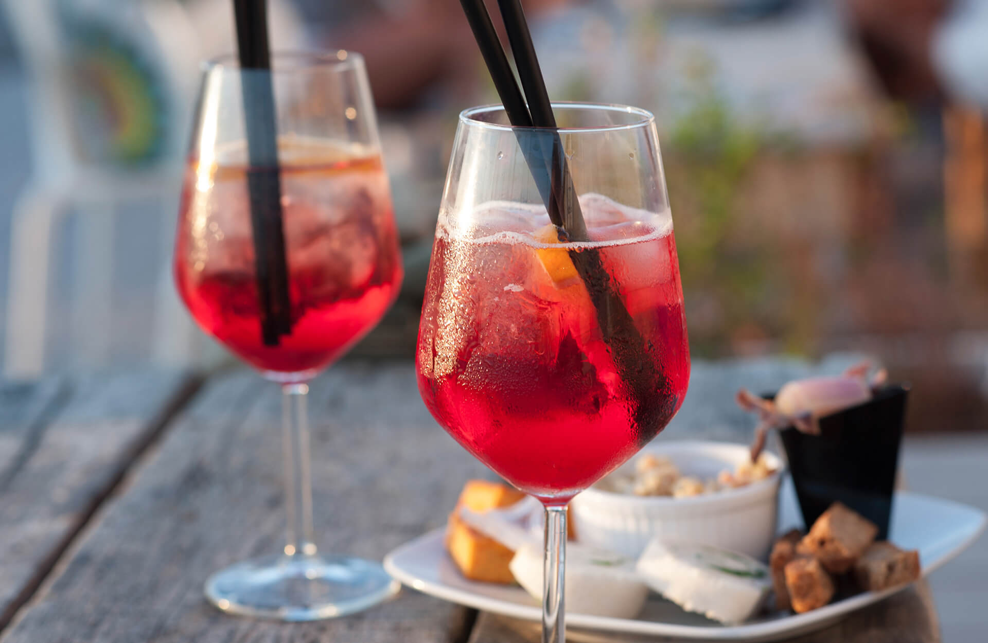 Two stemmed glasses filled with a red iced cocktail, garnished with orange slices and black straws, invite you into the world of taste as they rest on a wooden table beside a plate of assorted snacks. The softly blurred background sets the mood.