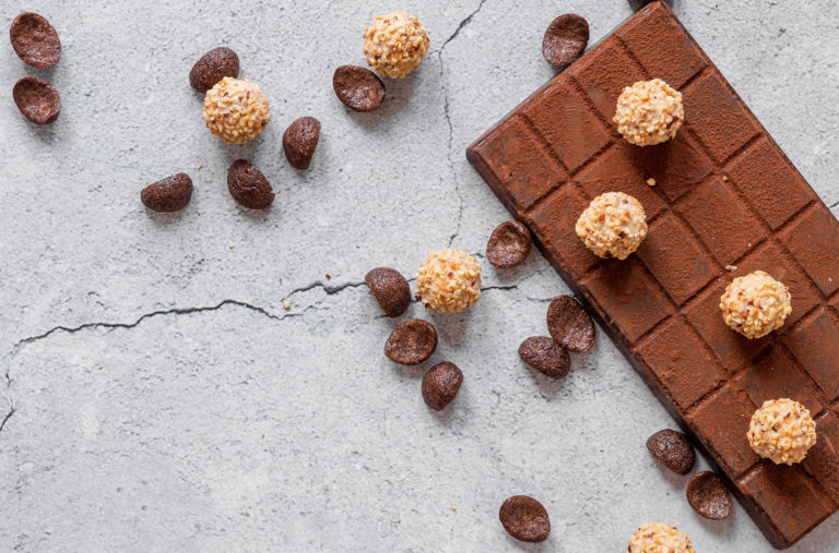 A textured chocolate bar topped with chocolate truffles is placed on a cracked gray surface. Chocolate chips are scattered around, adding to the arrangement's aesthetic.