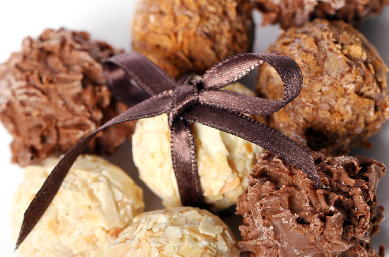 A close-up of assorted chocolate truffles with various coatings, including milk, white chocolate, and nuts. The truffles are tied together with a dark brown ribbon, creating an elegant presentation.