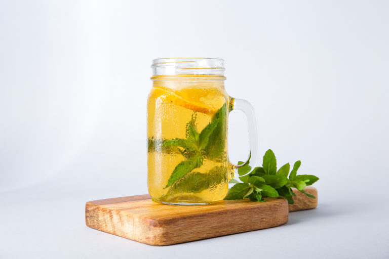 A mason jar filled with refreshing iced tea, a delightful nonalcoholic drink, is garnished with lemon slices and mint leaves on a wooden board. Fresh mint leaves rest beside the jar, set against a plain, light gray background.