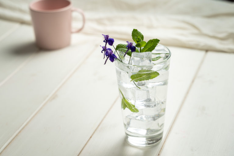 A tall glass of nonalcoholic drinks with ice, mint leaves, and small purple flowers sits on a white wooden table. In the blurred background, there's a pink mug. The scene is softly lit, creating a fresh and calm atmosphere.