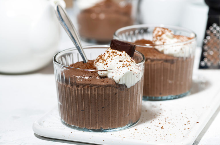 Two glass cups filled with chocolate mousse are topped with whipped cream and a piece of dark chocolate. A spoon rests inside one cup. Cocoa powder is sprinkled on top and around the cups, with a white teapot blurred in the background.