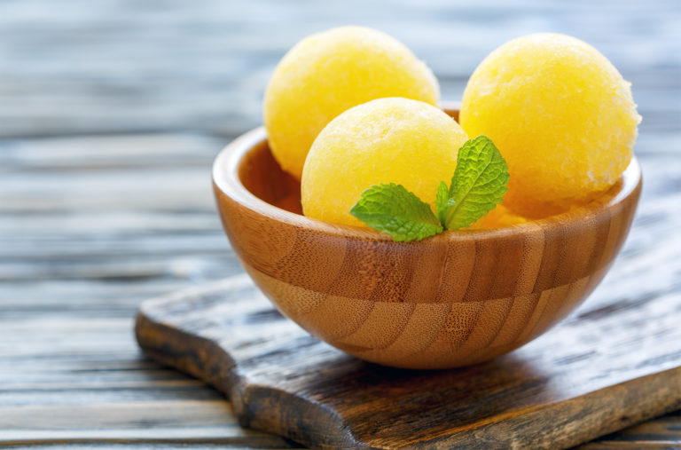 A wooden bowl holds three scoops of yellow sorbet, garnished with a sprig of mint. The bowl rests on a wooden surface with a weathered texture in the background, creating a fresh and inviting scene.