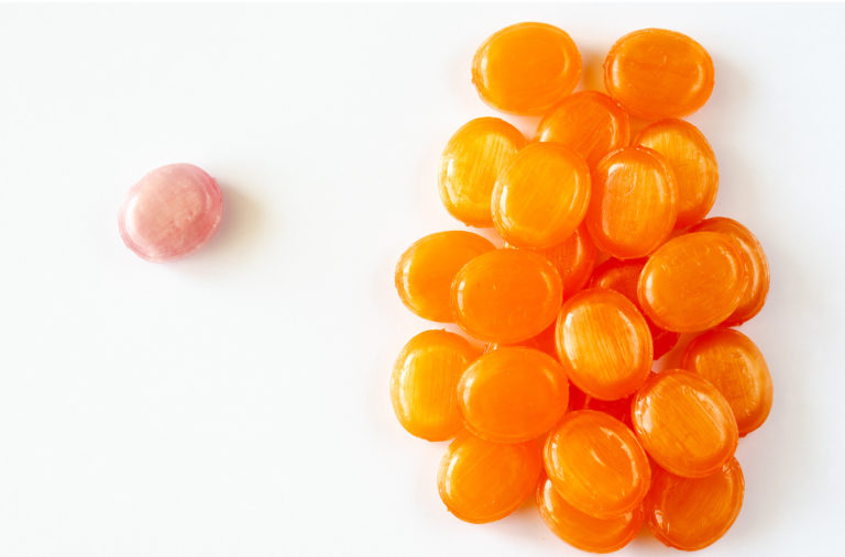 A single pink candy is separated from a cluster of twenty-six orange candies on a white background. The orange candies are grouped closely on the right, with the pink candy alone on the left, creating a contrast in color and arrangement.