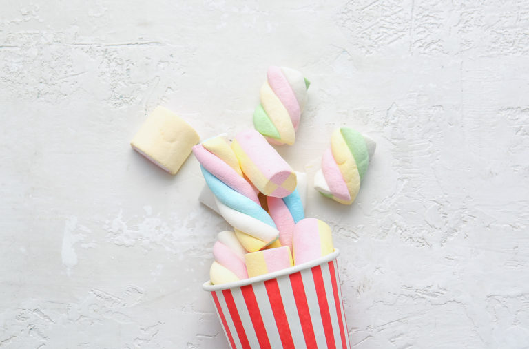 A red and white striped cup is tipped over, spilling pastel-colored marshmallows onto a textured white surface. The marshmallows are a mix of twisted and cylindrical shapes in soft hues of pink, blue, yellow, and green.