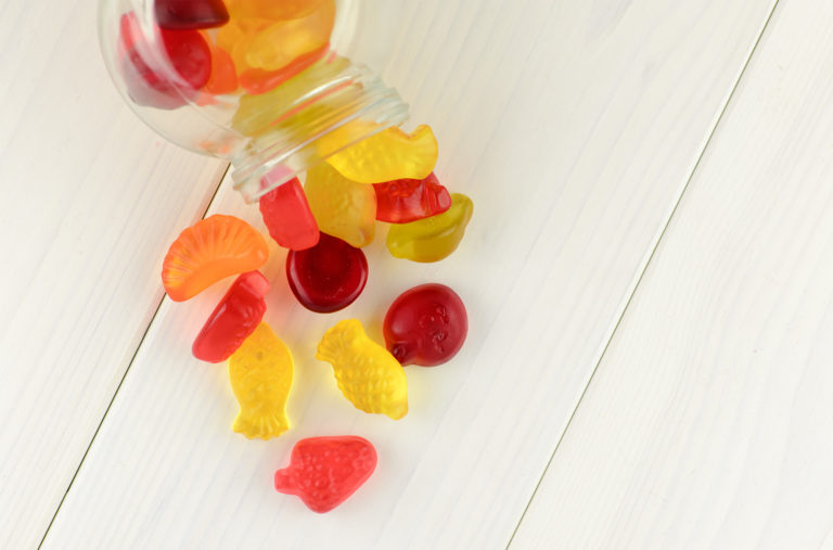 A glass jar tipped over on a white wooden surface, spilling colorful gummy candies. The candies are shaped like fruits and sea creatures, in red, yellow, and orange hues.
