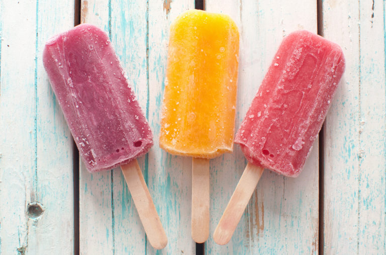 Three colorful popsicles sit on a rustic, white wooden surface. From left to right, the popsicles are purple, yellow, and red. Each has visible frost, indicating they are frozen.