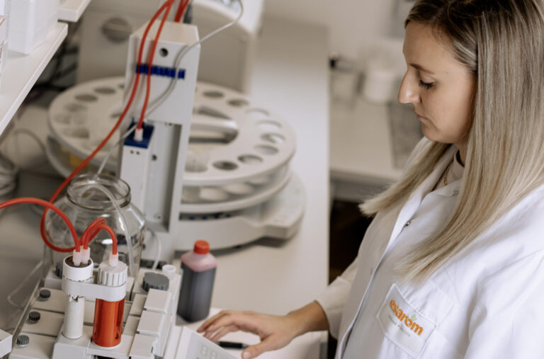 A woman in a lab coat is working in a laboratory. She stands beside scientific equipment with red tubes and a circular device. The setting includes shelves and various bottles and containers.