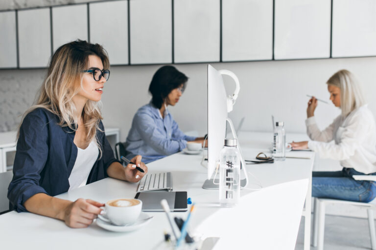 Trois femmes travaillent à un long bureau blanc dans un bureau. La femme au premier plan, portant des lunettes, tient un stylo et une tasse de café. À l'arrière-plan, deux femmes sont concentrées sur leurs ordinateurs, des bouteilles d'eau à proximité.