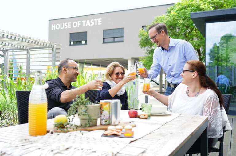 Four people are sitting at a table outdoors, toasting with glasses of juice. The table is set with snacks, a pitcher of juice, and decorative plants. They smile under a clear sky beside a building labeled "HOUSE OF TASTE.