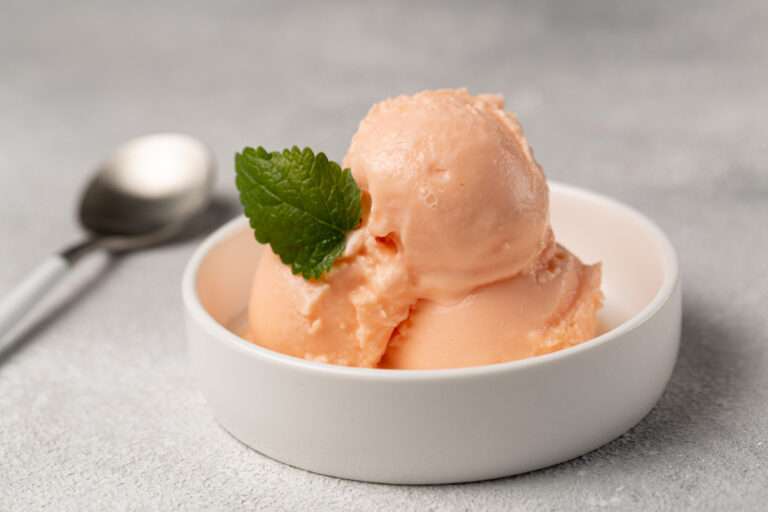 Two scoops of flavoured peach colored ice cream are in a white bowl, garnished with a small green mint leaf. A spoon lies on the gray surface next to the bowl.