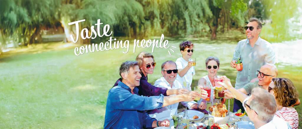 A group of people sitting at an outdoor table in a park, raising glasses filled with refreshing beverages in a toast. They appear to be enjoying a picnic. Text overlay reads, "Taste connecting people." The background features lush greenery and a sunny atmosphere.