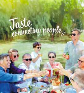 A group of people enjoying a picnic in a sunny park, holding beverages and more, smiling at each other. A large text overlay reads, "Taste connecting people." The table is set with delicious ingredients and is surrounded by lush greenery.