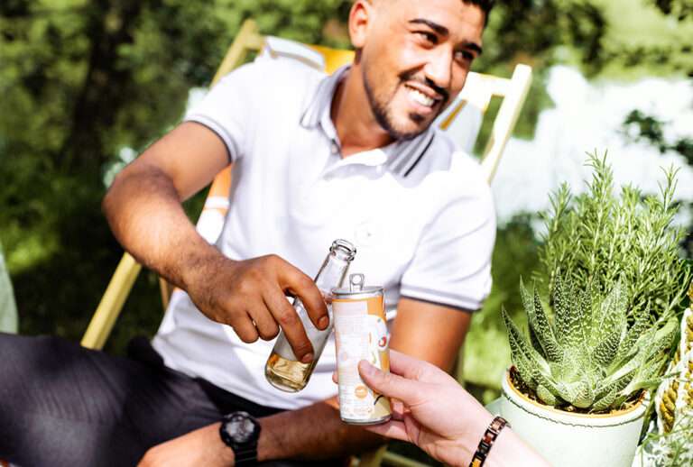 A man in a white shirt smiles while clinking a tasty glass bottle with another person's energy drink in an outdoor setting. They are sitting on chairs, with greenery and a potted plant in the foreground.