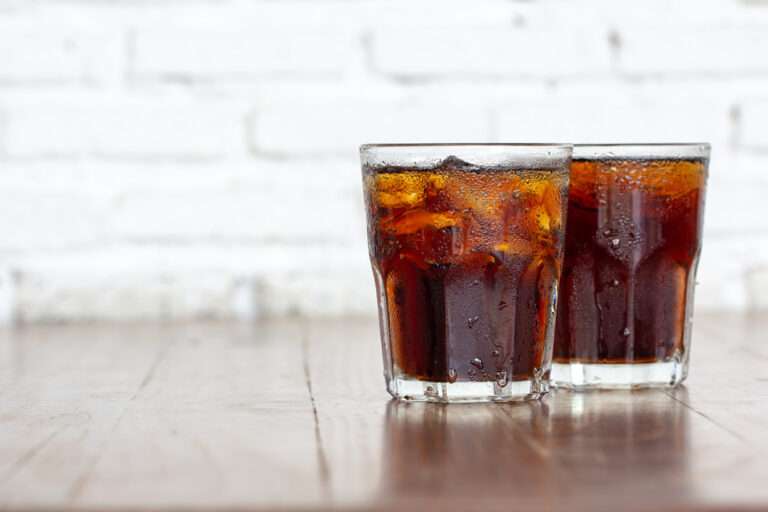 Two glasses of iced cola sit on a wooden table against a white brick wall. The glasses are filled with ice cubes and the cola appears chilled, with condensation visible on the glass surfaces.