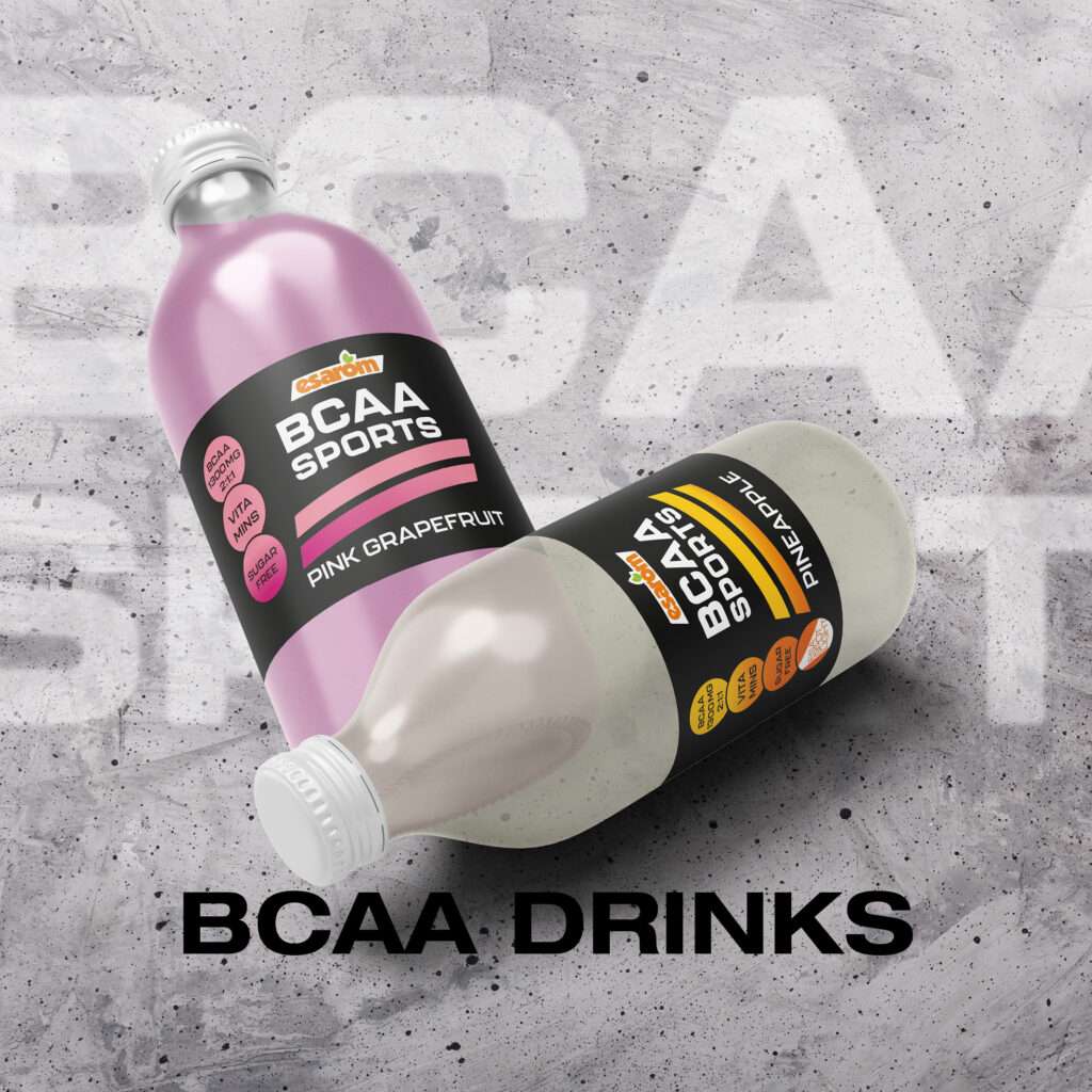 Two BCAA SPORTS drinks bottles, one pink grapefruit and one pineapple, lie on a textured gray surface with "BCAA DRINKS" below them. Both bottles, perfect for prosweets fans, have black labels with colorful accents.