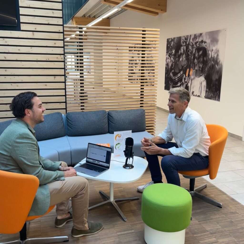 Zwei Männer sitzen sich in einer modernen Bürolounge gegenüber und führen ein Gespräch. Ein Laptop und ein Mikrofon stehen auf dem Tisch zwischen ihnen. Die Umgebung ist mit bunten Stühlen, einer hölzernen Trennwand und Wandkunst ausgestattet.
