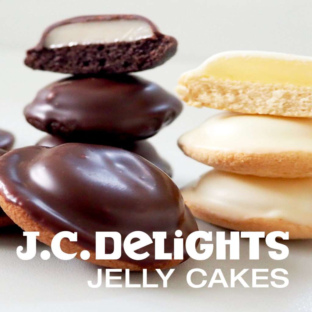 A close up of chocolate and vanilla covered jelly cakes, some stacked and one cut open to reveal a jelly layer inside. The text reads “J.C. Delights Jelly Cakes.”.