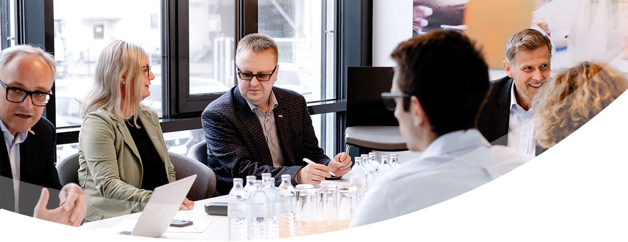 A group of professionals sit around a conference table engaged in discussion at Essenzfabrik, founded by esarom, with laptops, documents, and water bottles in front of them. Large windows in the background let in natural light.