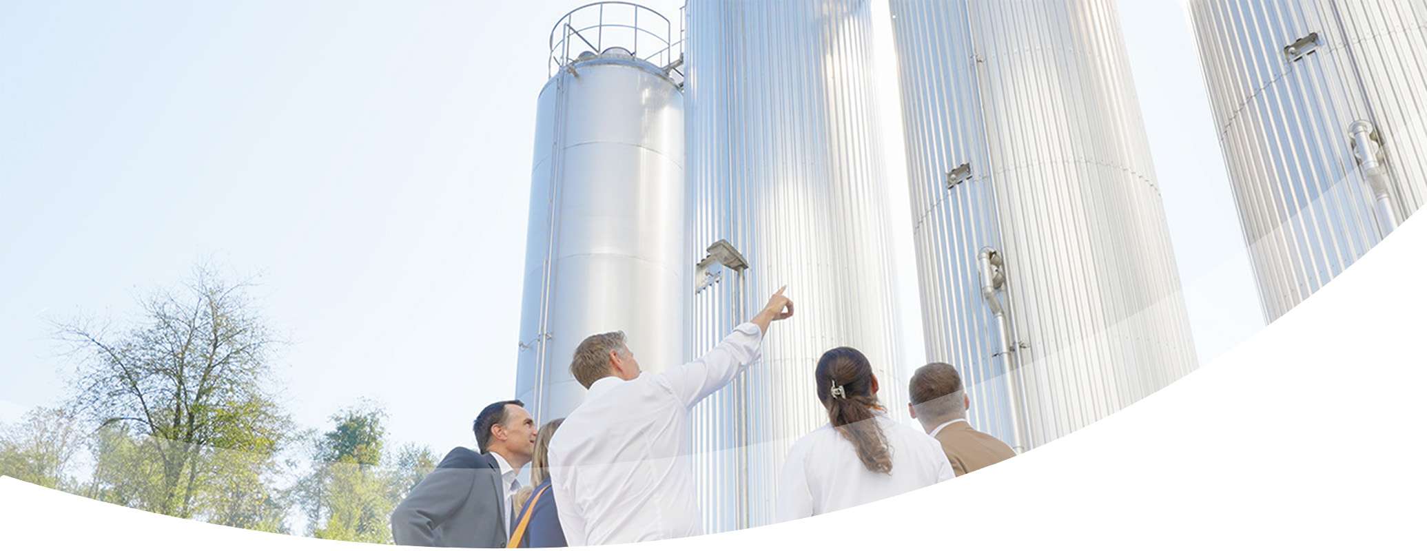 Four people in business attire stand outdoors, discovering tall, cylindrical industrial tanks under a clear sky, with trees in the background—an experience that sparks curiosity and insight.