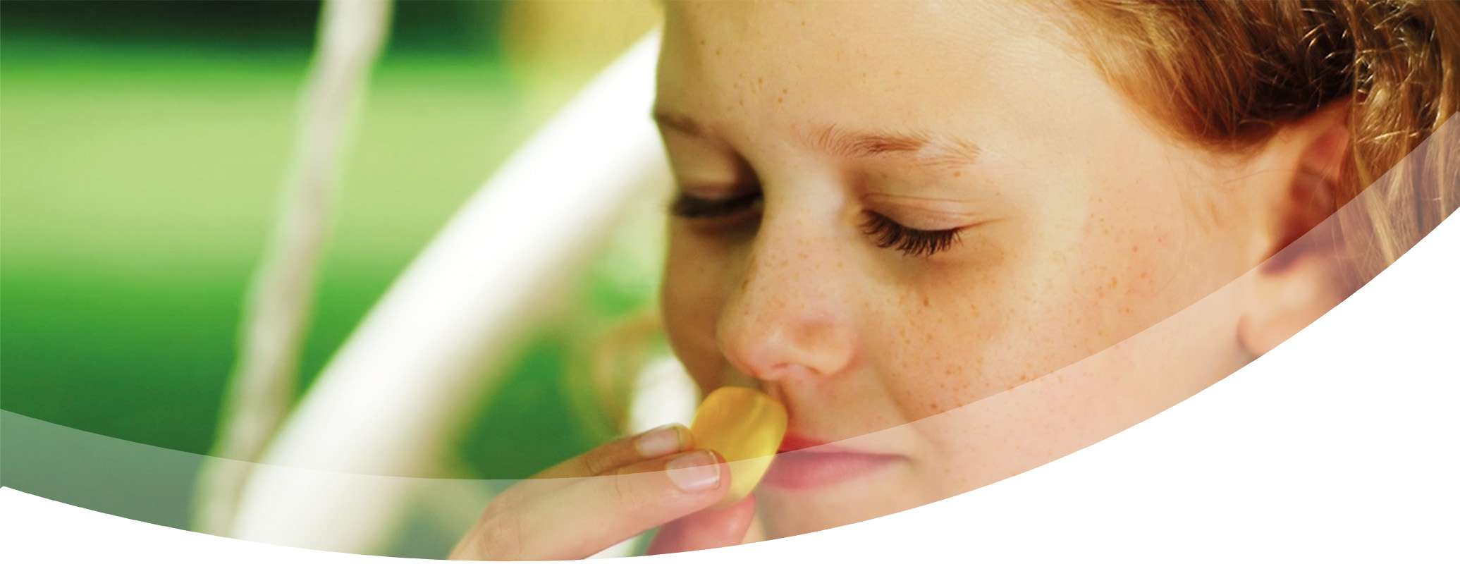A close up of a young child with freckles and curly hair, eyes closed, smelling a yellow flower petal, bathed in gentle lighting and a soft green background—capturing a moment as sweet as confectionery.