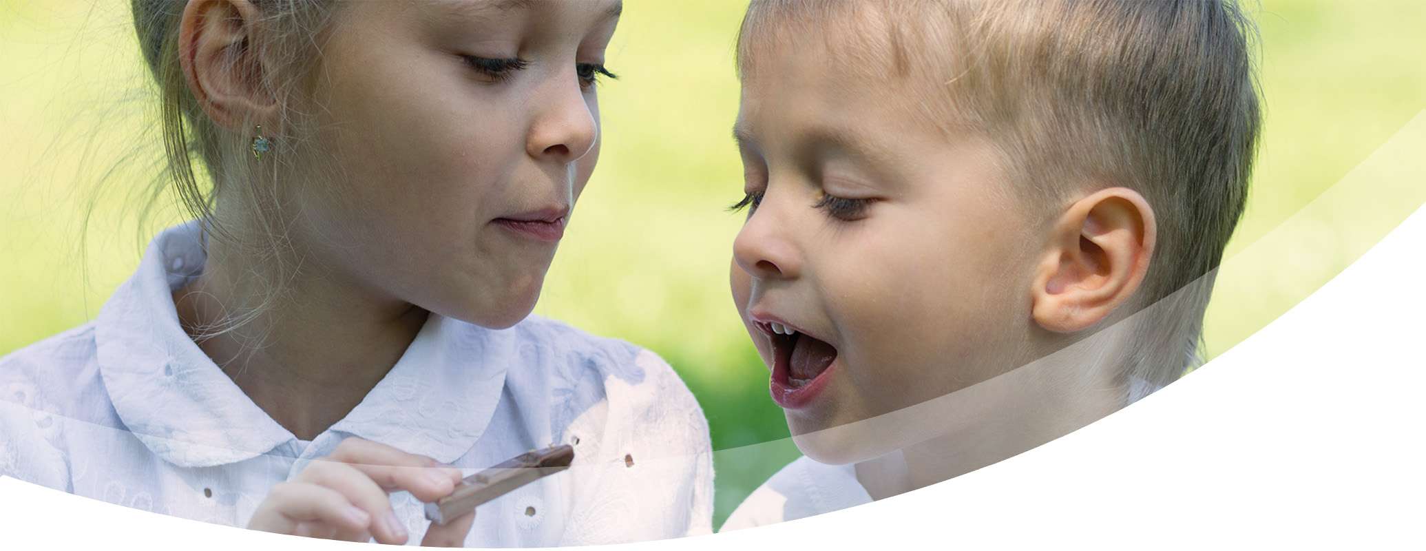 Two young children in white shirts sit outdoors. One child holds a wooden spoon with chocolate, feeding it to the other, who eagerly awaits the treat with eyes closed. The background is lush and green, softly blurred.