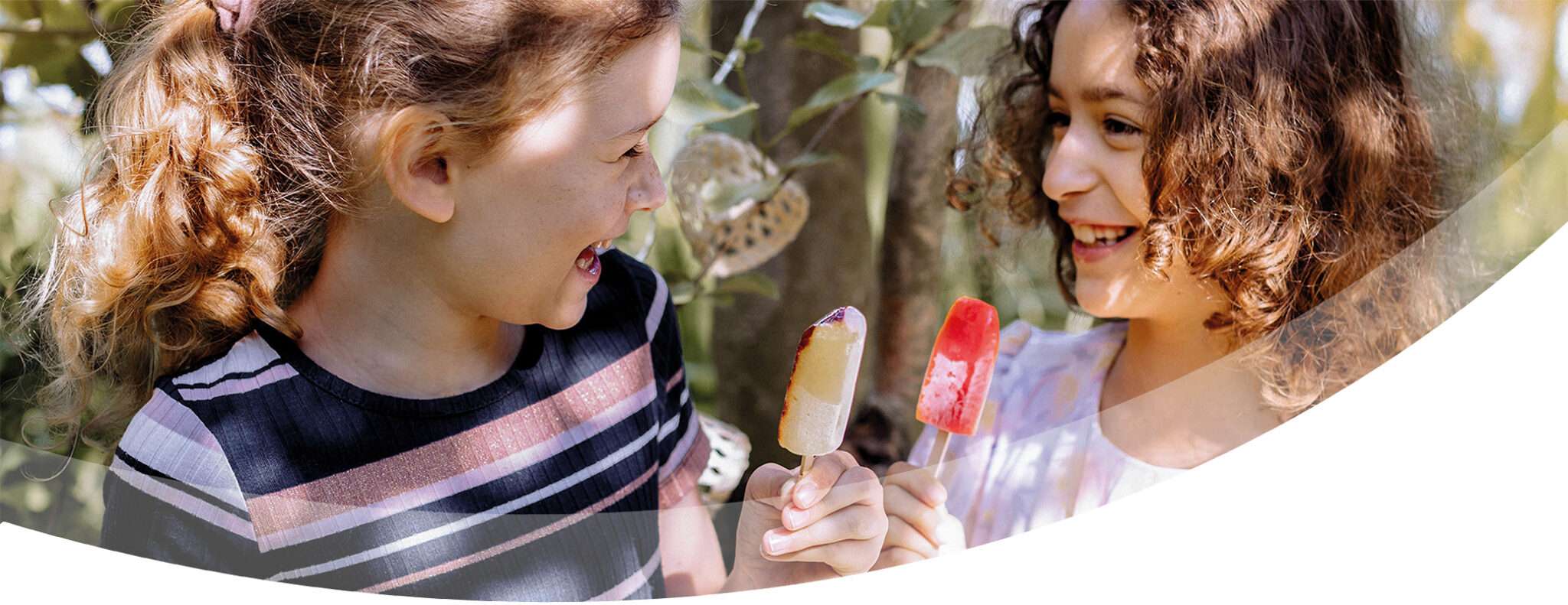 Two young girls with curly hair smile at each other while holding and enjoying colorful popsicles and ice cream outdoors, surrounded by greenery and sunlight.