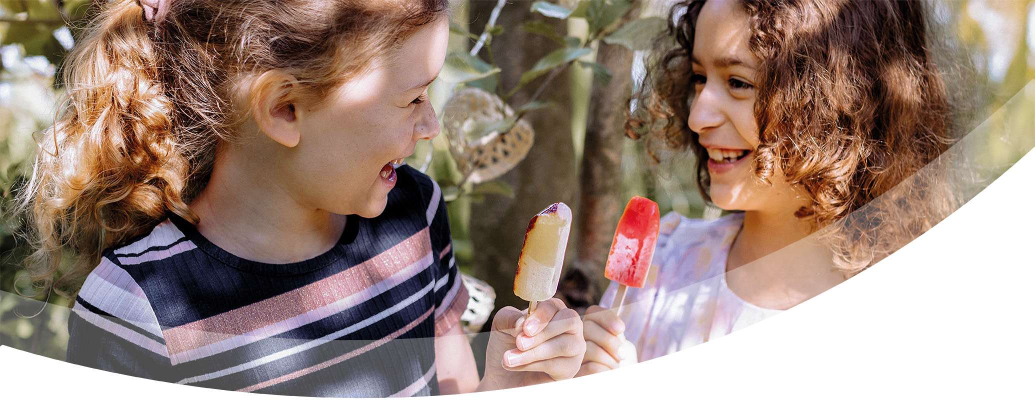 Two young girls with curly hair smile at each other while holding and enjoying colorful popsicles and ice cream outdoors, surrounded by greenery and sunlight.