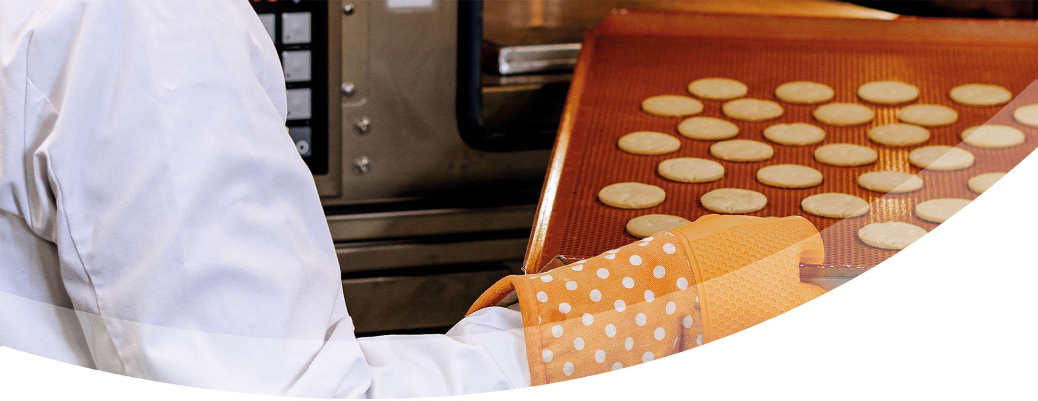 A person in a white coat uses orange oven mitts with white dots to hold a tray of round pastry cookies near an oven.