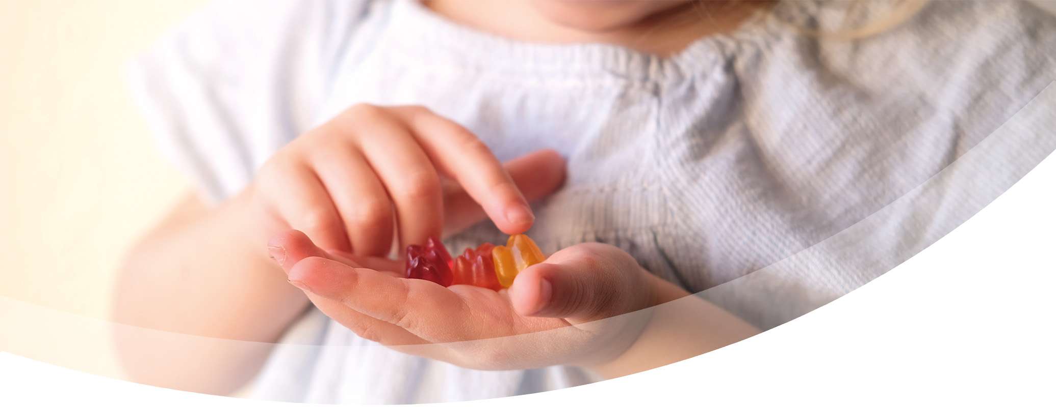 A young child in a light blue shirt holds colorful, sugar coated gummy candies in one hand while picking one up with the other. The image is softly focused on the hands and candies.