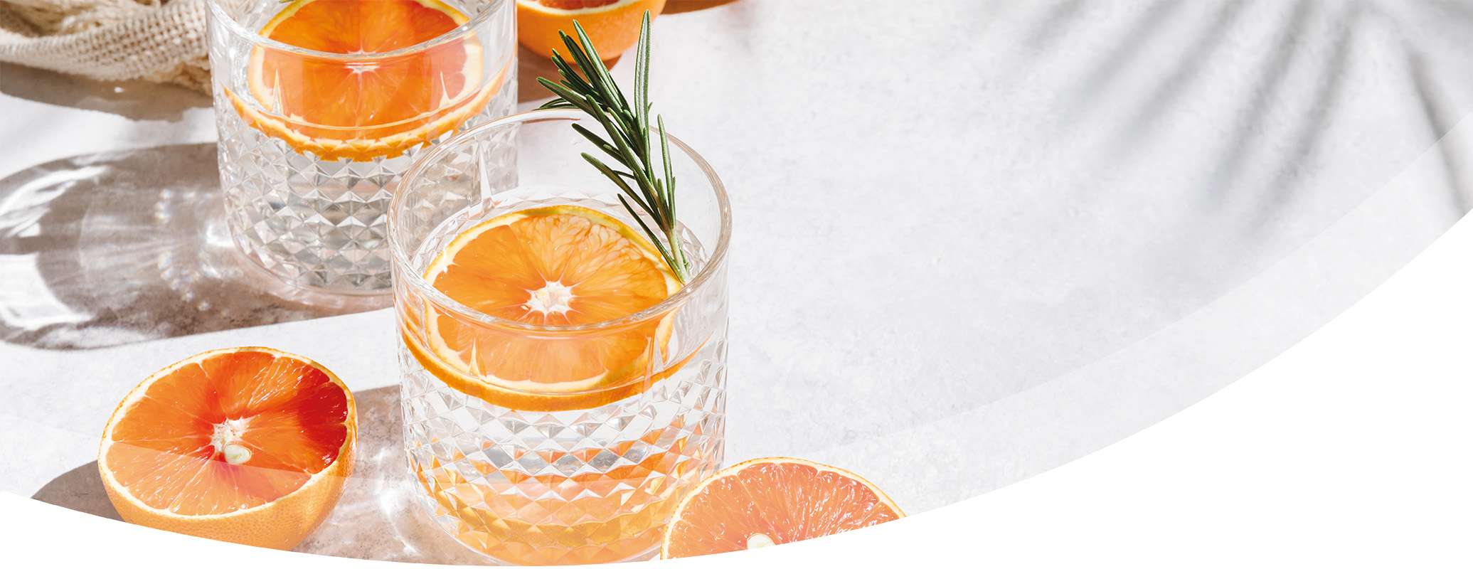Two crystal glasses filled with an alcoholic clear drink, garnished with orange slices and a rosemary sprig, sit on a light surface with halved oranges nearby and dappled sunlight casting soft shadows.