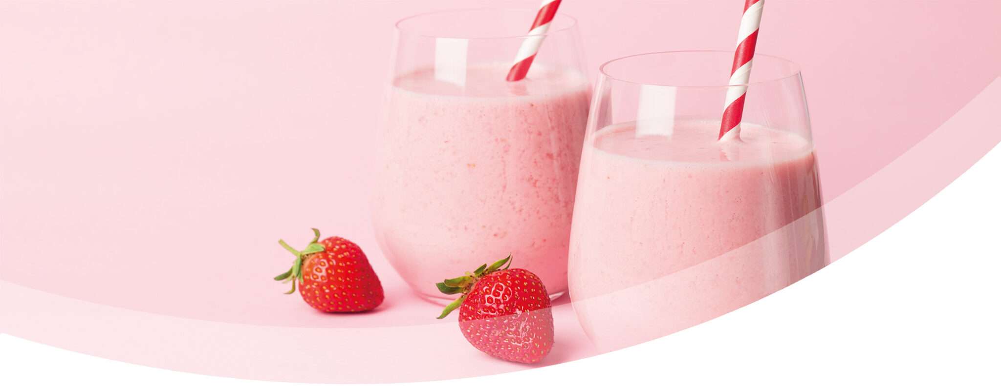 Two glasses of creamy, dairy strawberry smoothie with red and white striped straws sit on a light pink surface, accompanied by two fresh strawberries.