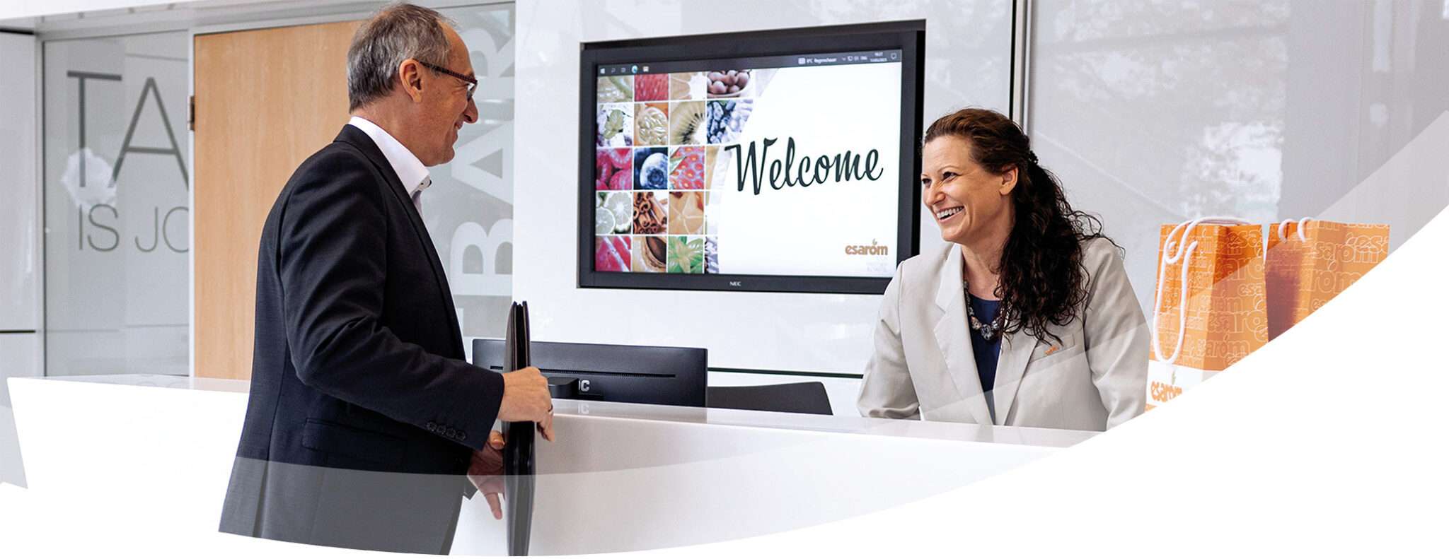 A man in a suit stands at a modern reception desk, smiling at a woman receptionist who is also smiling. A welcome sign and colorful images promoting business opportunities are displayed on a screen behind the desk.