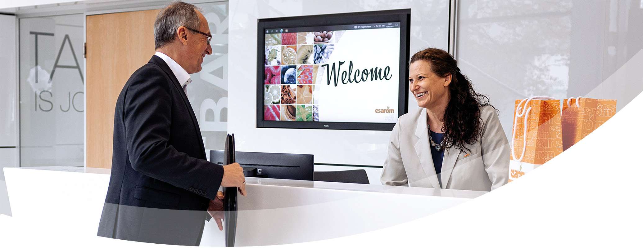 A man in a suit stands at a modern reception desk, smiling at a woman receptionist who is also smiling. A welcome sign and colorful images promoting business opportunities are displayed on a screen behind the desk.