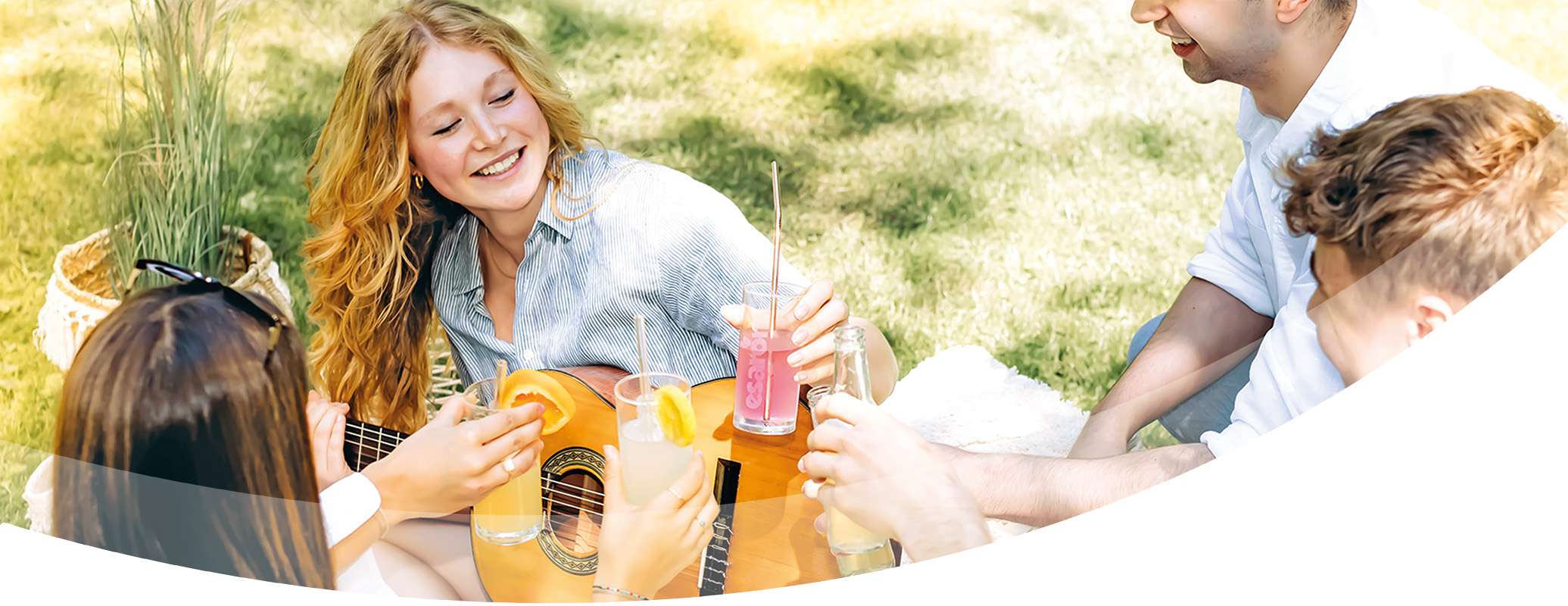 A group of young adults sit on the grass, enjoying a picnic. One person plays a guitar while others hold colorful drinks and smile at each other in the sunlight.
