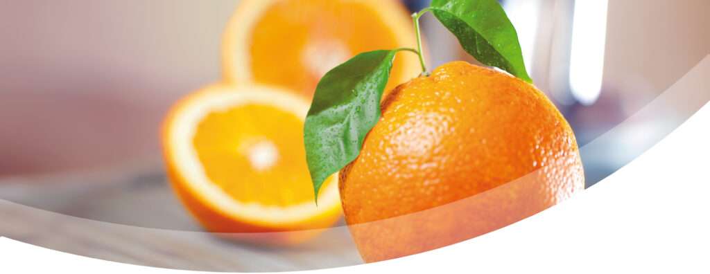 A close up of a fresh orange with green leaves, with a sliced orange in the background, all set on a light surface with a soft, blurred backdrop.