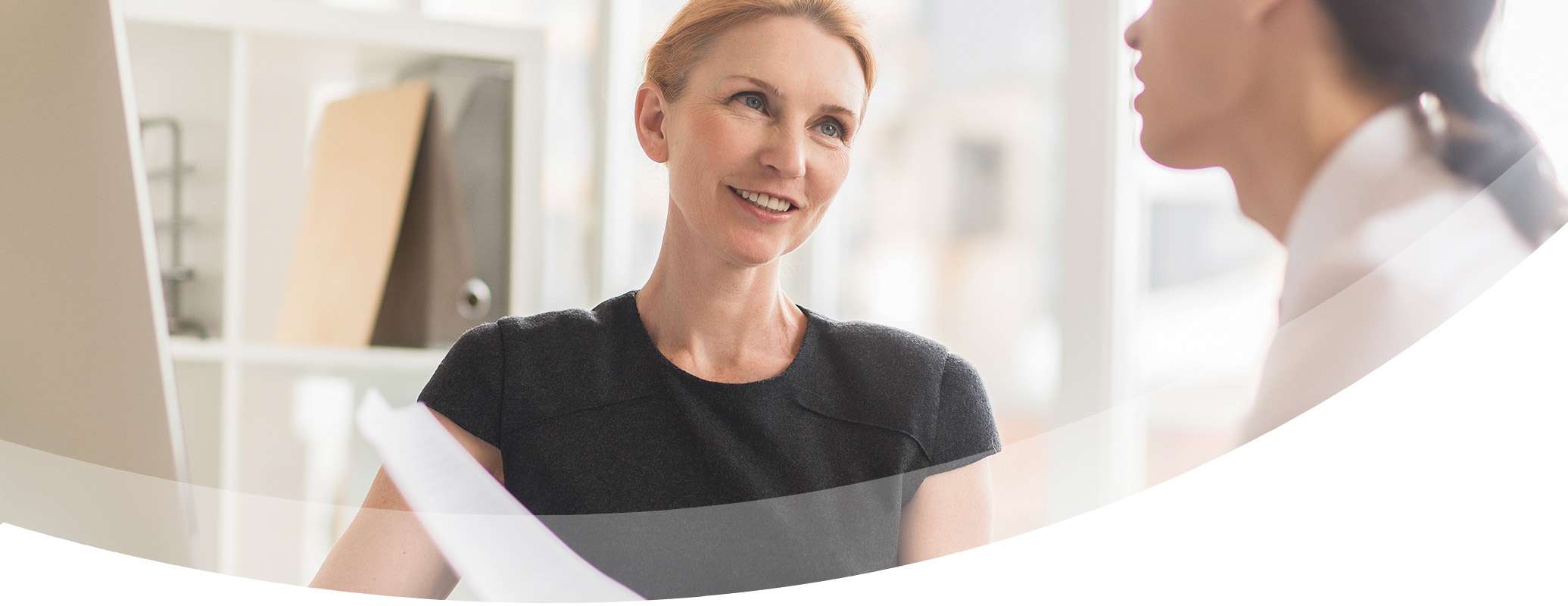 A woman with red hair smiles while holding papers, sitting at a desk and talking to another person in an office setting with shelves in the background.