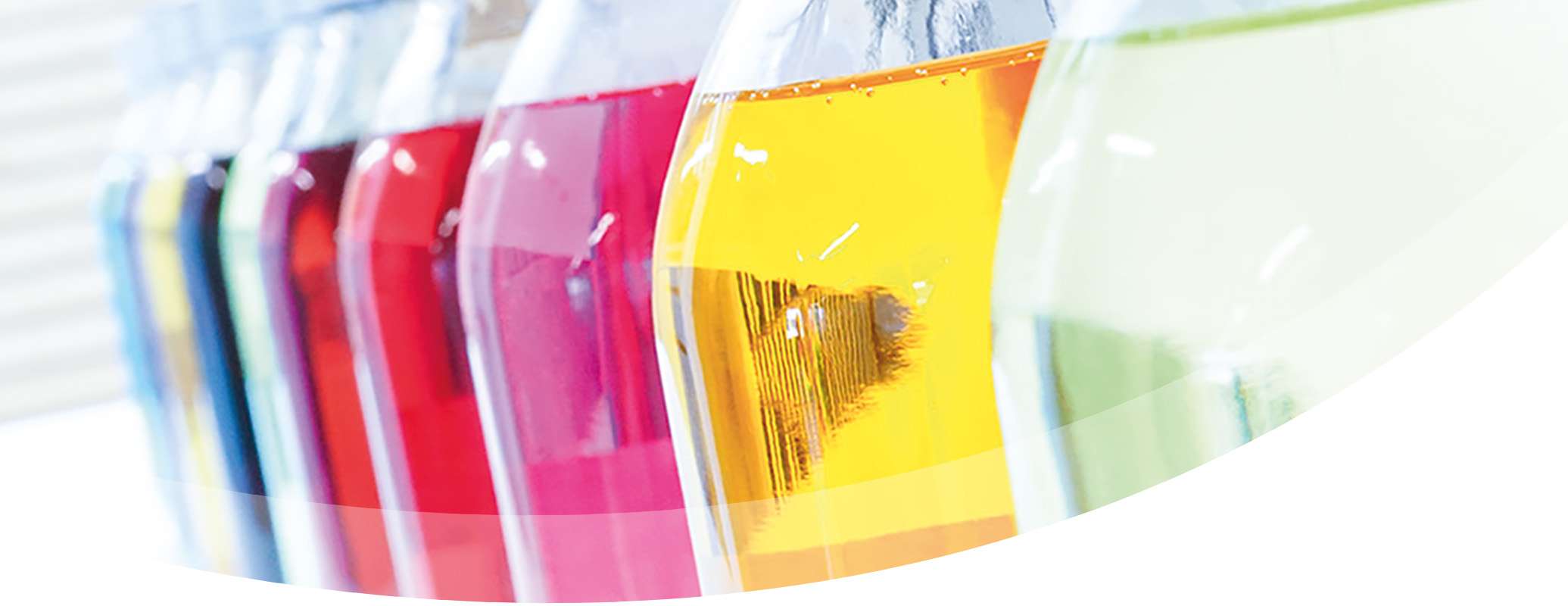 Close up of several glass bottles filled with colourful liquids, each showcasing a different flavour, arranged in a row on a white surface with a bright, blurred background.