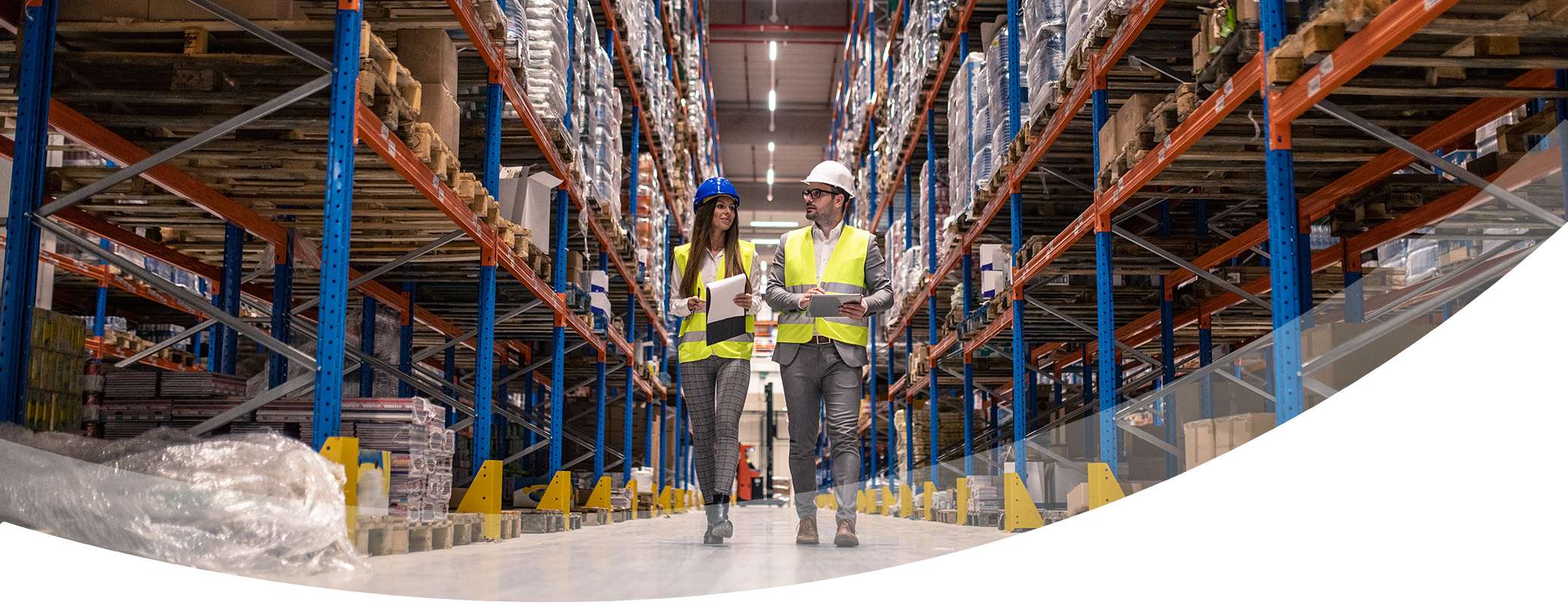 Two people in safety vests and helmets walk through a warehouse aisle lined with tall shelves full of boxes, discussing Produktionslogistik while holding clipboards and a tablet.