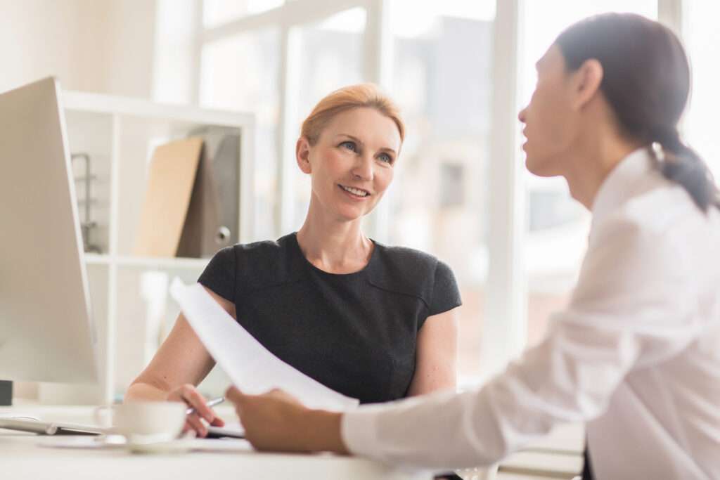 Two women in an office setting have a conversation at a desk. One woman, smiling, holds papers while listening to the other, who is speaking. Shelves and a computer are in the background.