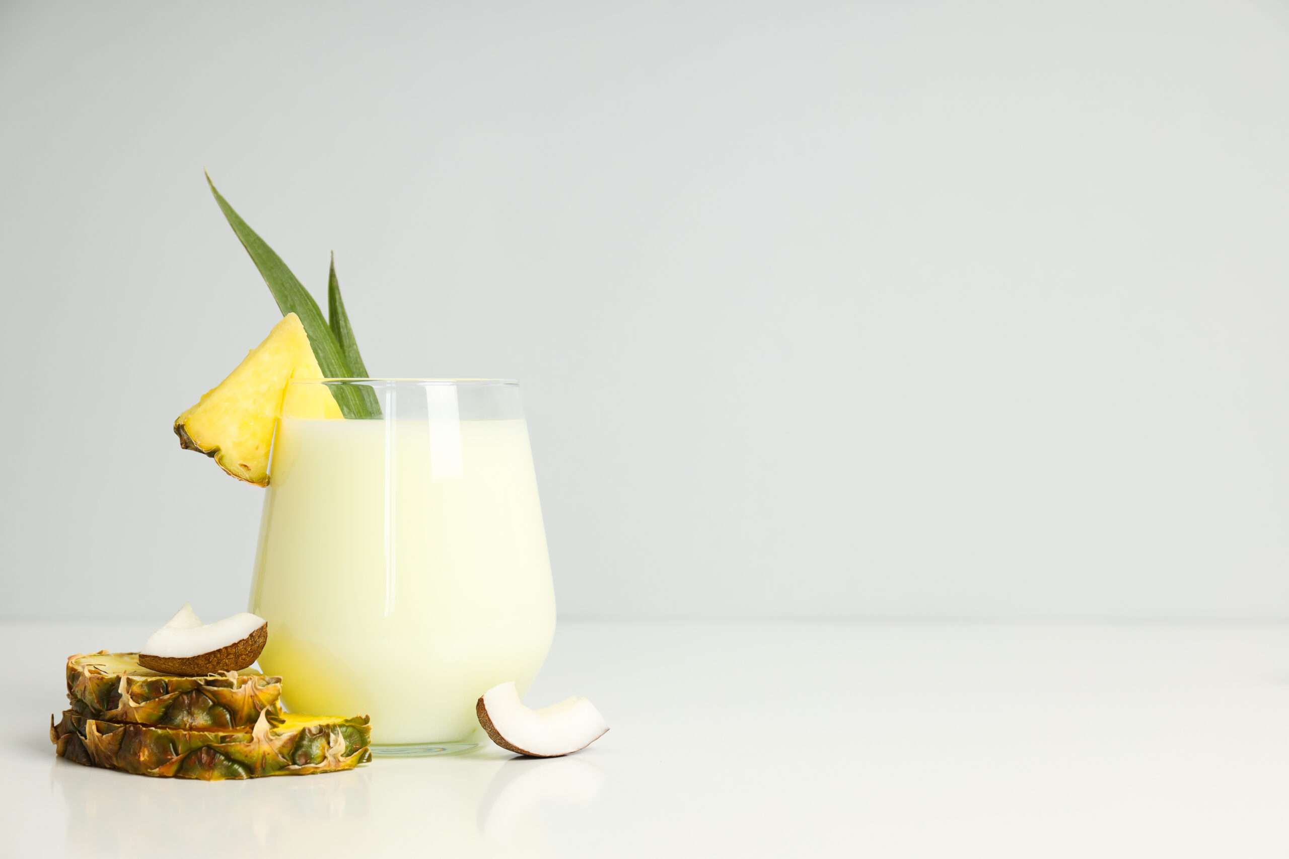 A glass of pineapple and coconut juice, garnished with a pineapple wedge and leaf, sits beside sliced pineapple and coconut on a white surface with a light gray background—perfect for pairing with refreshing Fibre Soda.