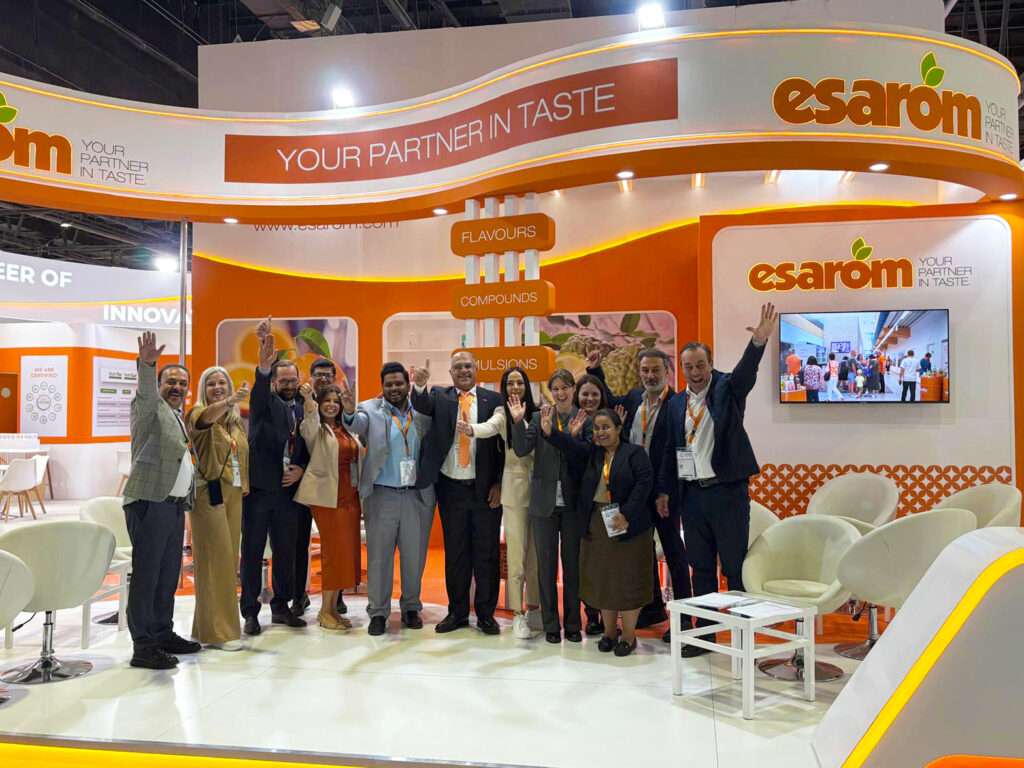 A group of people stand together, smiling and waving, in front of an orange and white exhibition booth with the esarom logo and the slogan "Your Partner in Taste." The booth features seating and product displays.