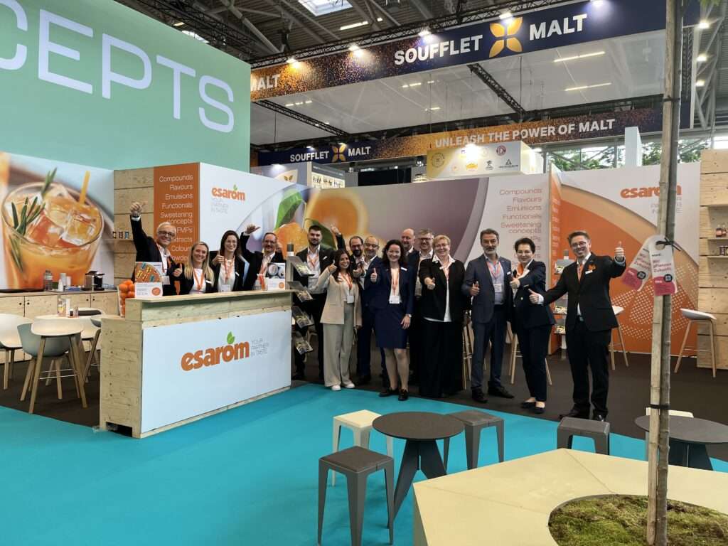 A group of people stand smiling and giving thumbs up at the esarom booth during a trade show, with a colorful display, banners, and a bar area in the background.