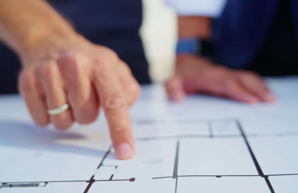 A close-up of a person's hand with a ring, pointing to a specific area on a floor plan or architectural blueprint, with another person nearby.