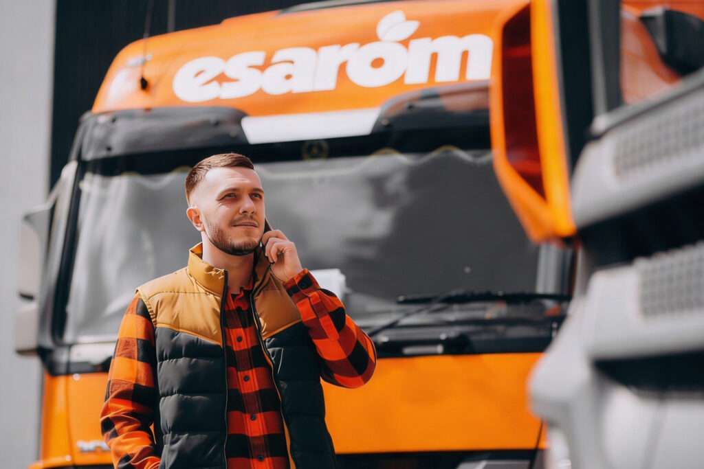 A man wearing a plaid shirt and vest stands in front of an orange truck, talking on his phone. The truck behind him has the word "esarom" visible on its front.