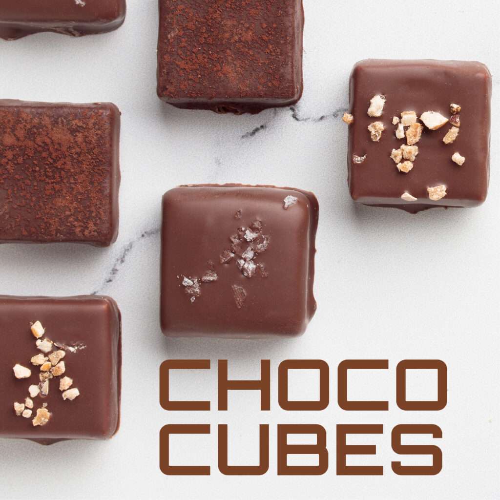 Five square chocolate cubes, some topped with salt or nut pieces, arranged on a light surface. The text "CHOCO CUBES" appears in bold brown letters at the bottom right.