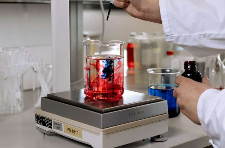 A person in a lab coat adds a dark liquid to a beaker of red liquid, creating swirling patterns, on a lab hot plate. Lab equipment and glassware are visible in the background.