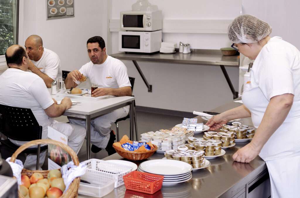 Four people in white uniforms are in a cafeteria; three are seated and eating at a table, while one is arranging food containers at a counter. Fruit and packaged food items are also visible on the counter.